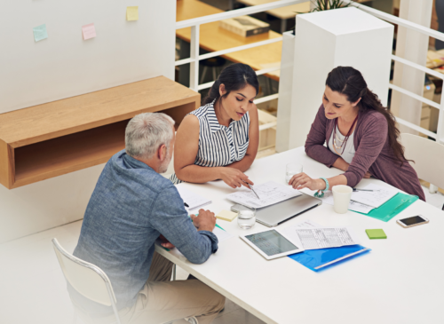 3 people sit at a table in a library looking at research documents.