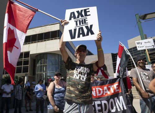 A protester holding a sign that says "Axe the Vax" outside with a crowd behind.