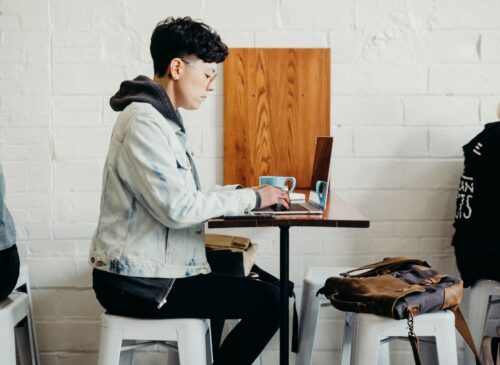 A person wearing a jean jacket sits at a cafe table working on a laptop.