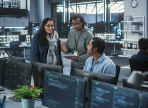 Three people stand together in a computer lab talking.